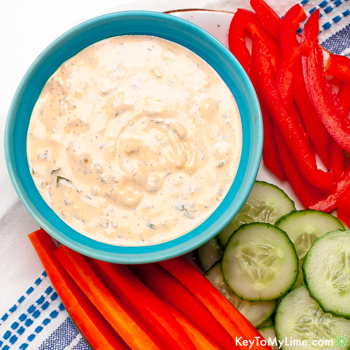 Spicy ranch in a small blue bowl surrounded by sliced carrots, cucumbers, and red bell pepper.