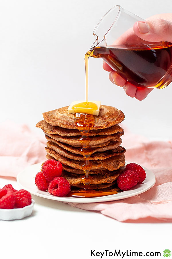A hand pouring syrup over a stack of oat flour pancakes.