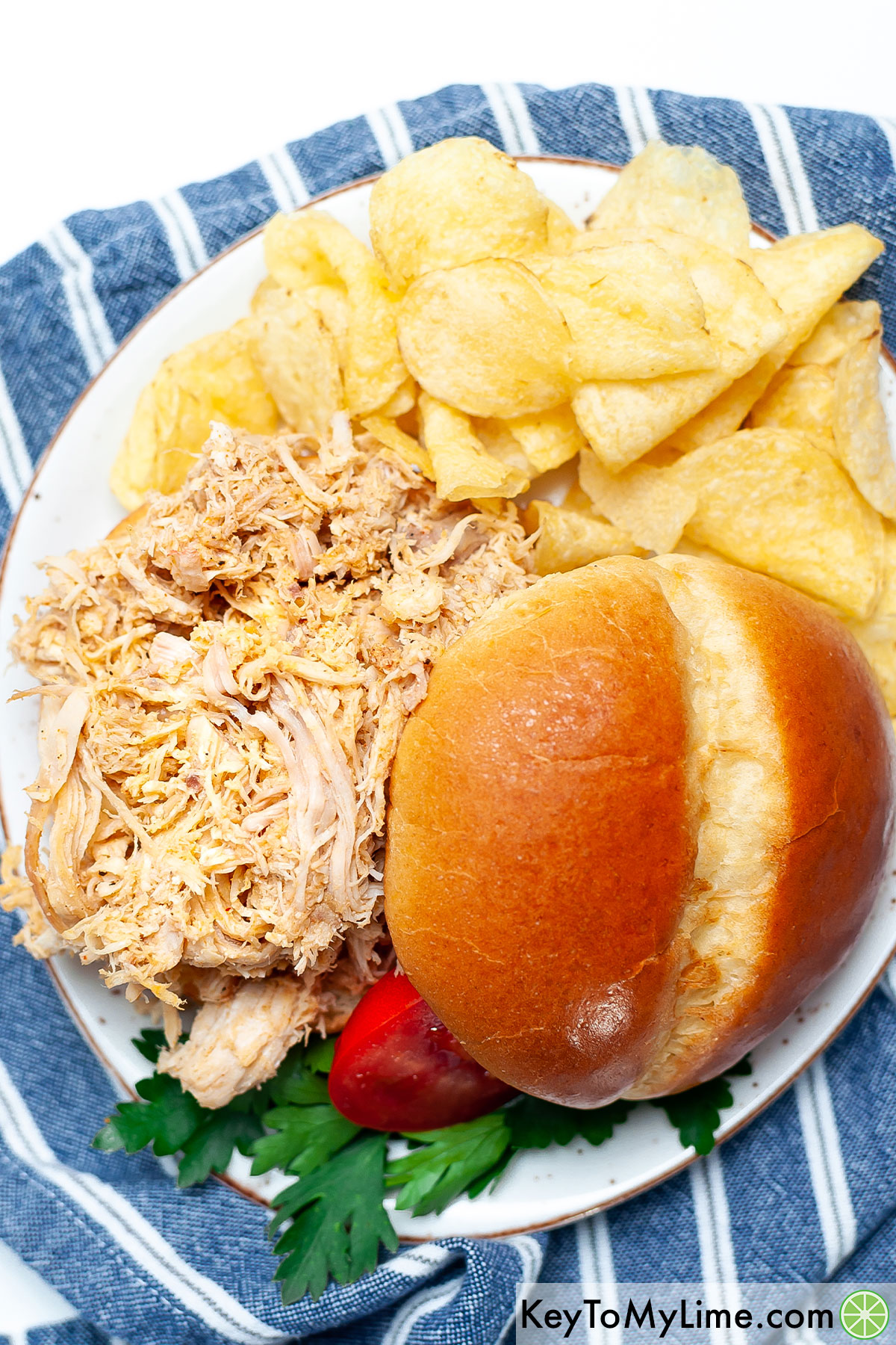 An overhead image of a pulled chicken sandwich on a plate with potato chips.