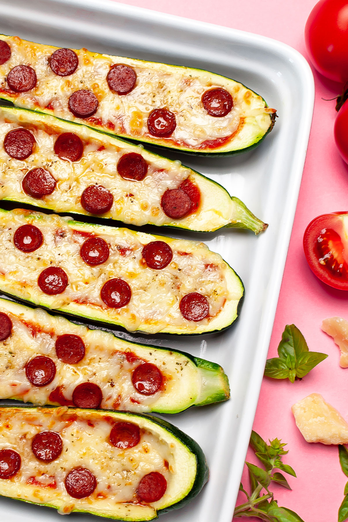 Zucchini pizza boats next to basil and tomato on a pink background.