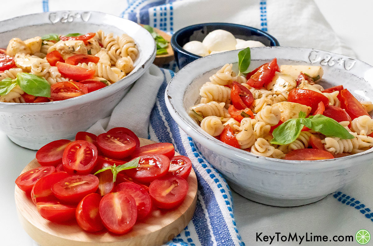 Two bowls of caprese pasta salad next to plates of mini mozzarella and sliced tomatoes.