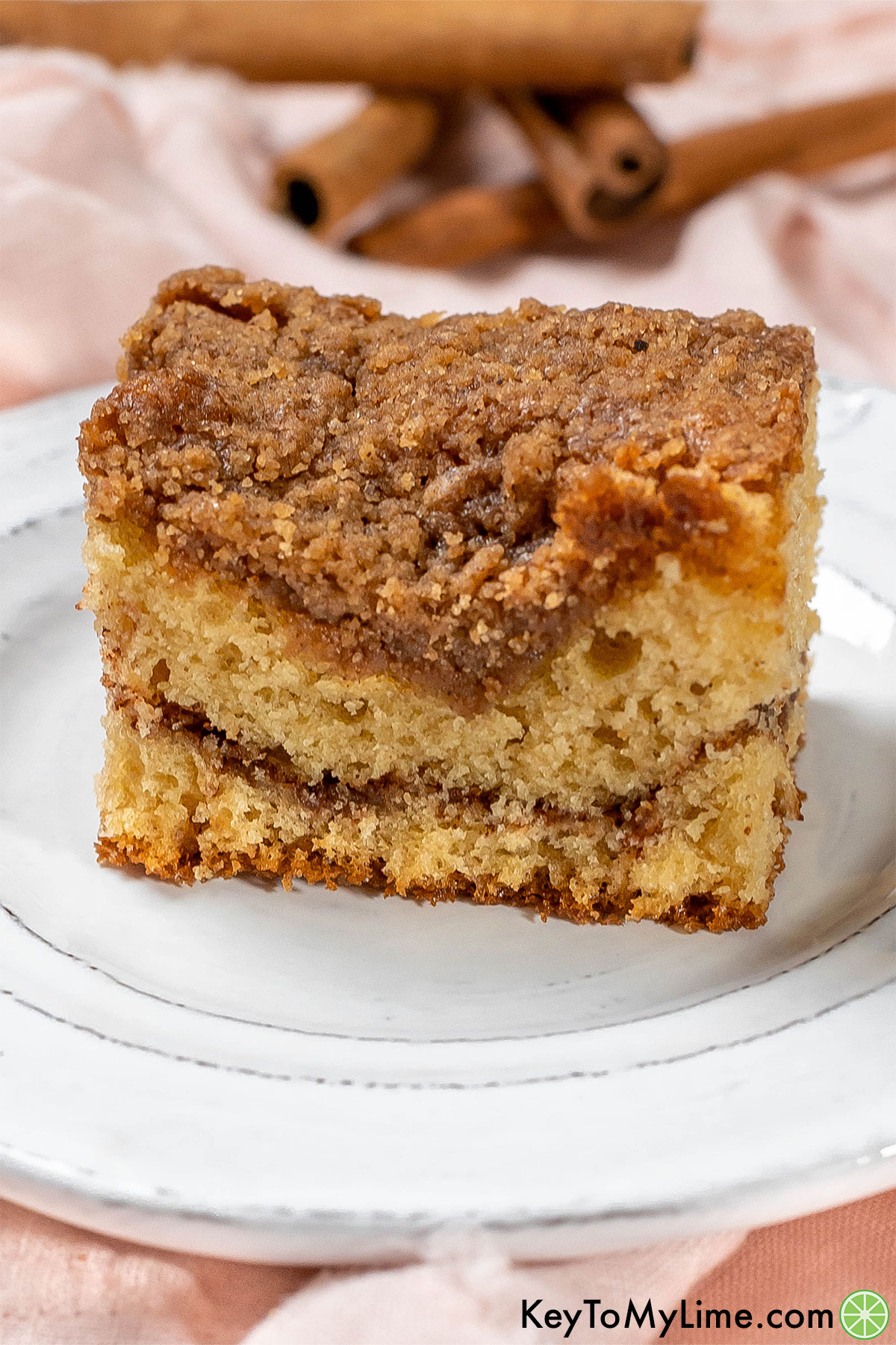 A close up image of a slice of coffee cake served on a white plate with cinnamon sticks in the background.