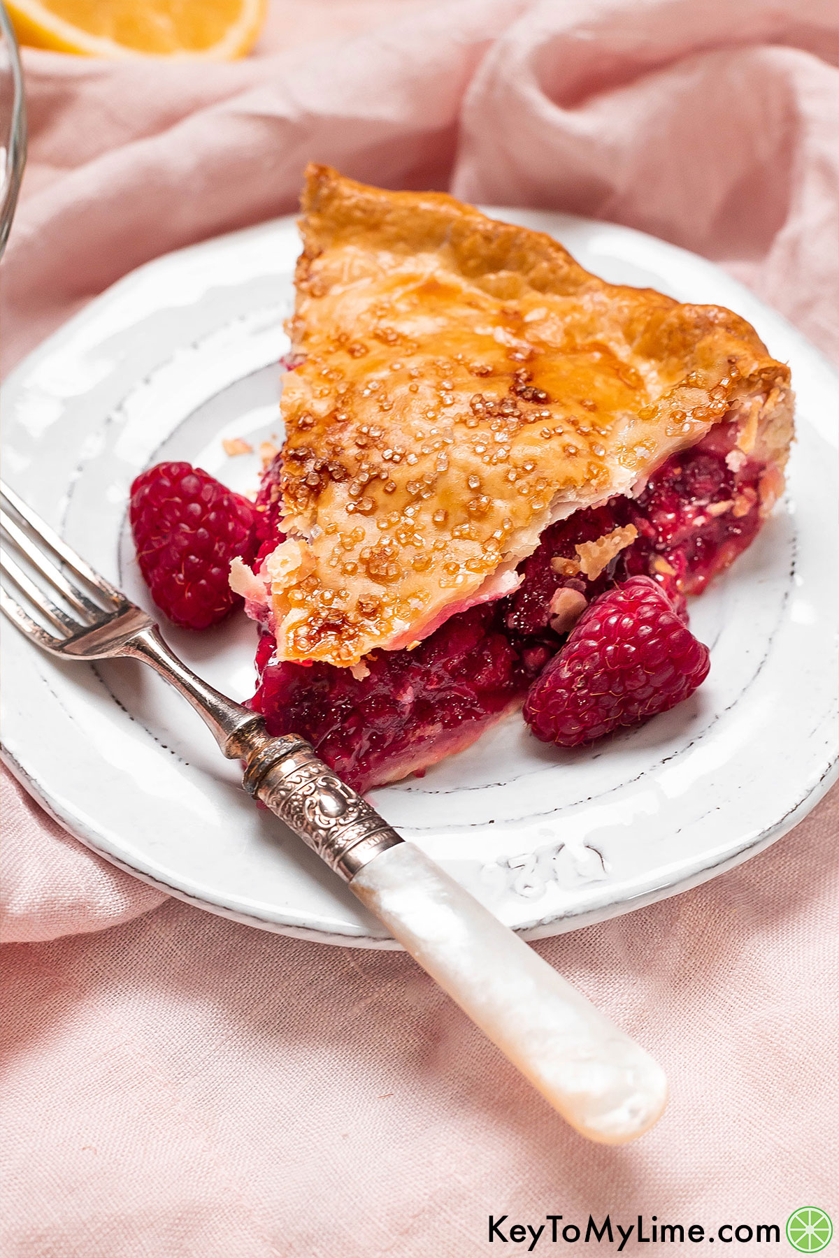 A slice of pie with a golden brown crust filled with a thick raspberry filling served on a white plate with fresh berries to the side.