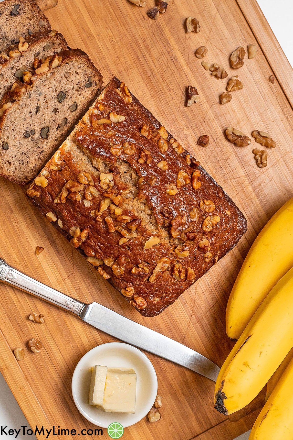 An overhead shot of glistening banana bread with a small dish of butter and a bundle bananas to the side.
