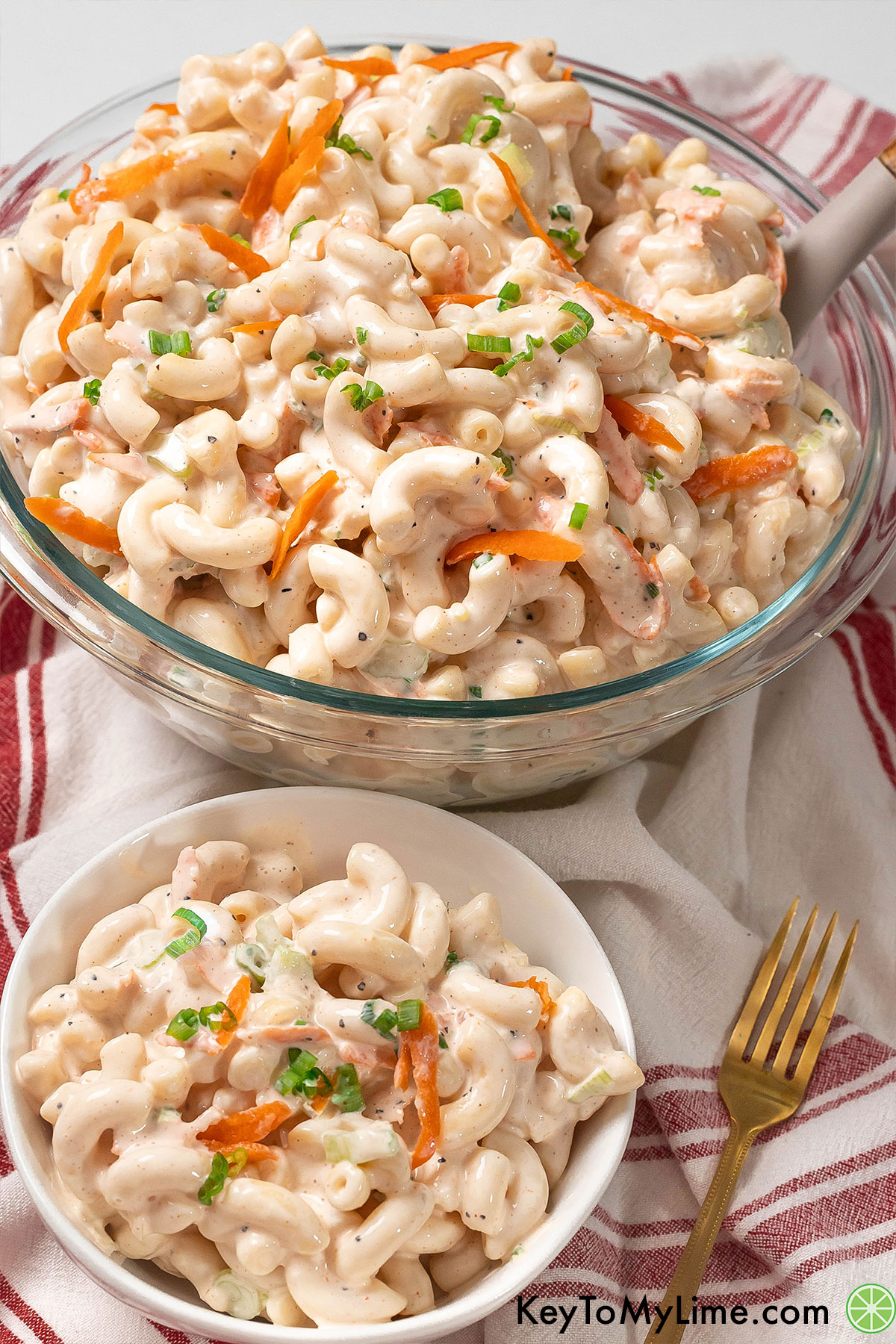 An angled shot of a large mound of flavorful macaroni salad in a glass ditch with carrots and green onion throughout.