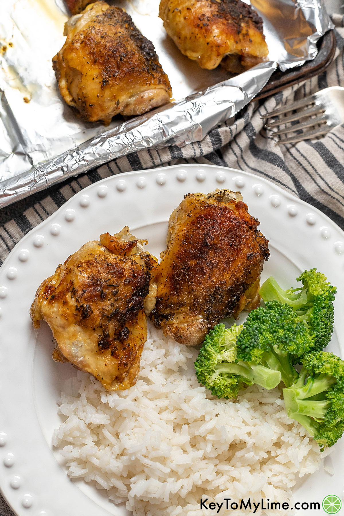 An overhead shot of golden crispy chicken thighs plated with freshly cooked broccoli and rice with thighs on baking sheets in the back.