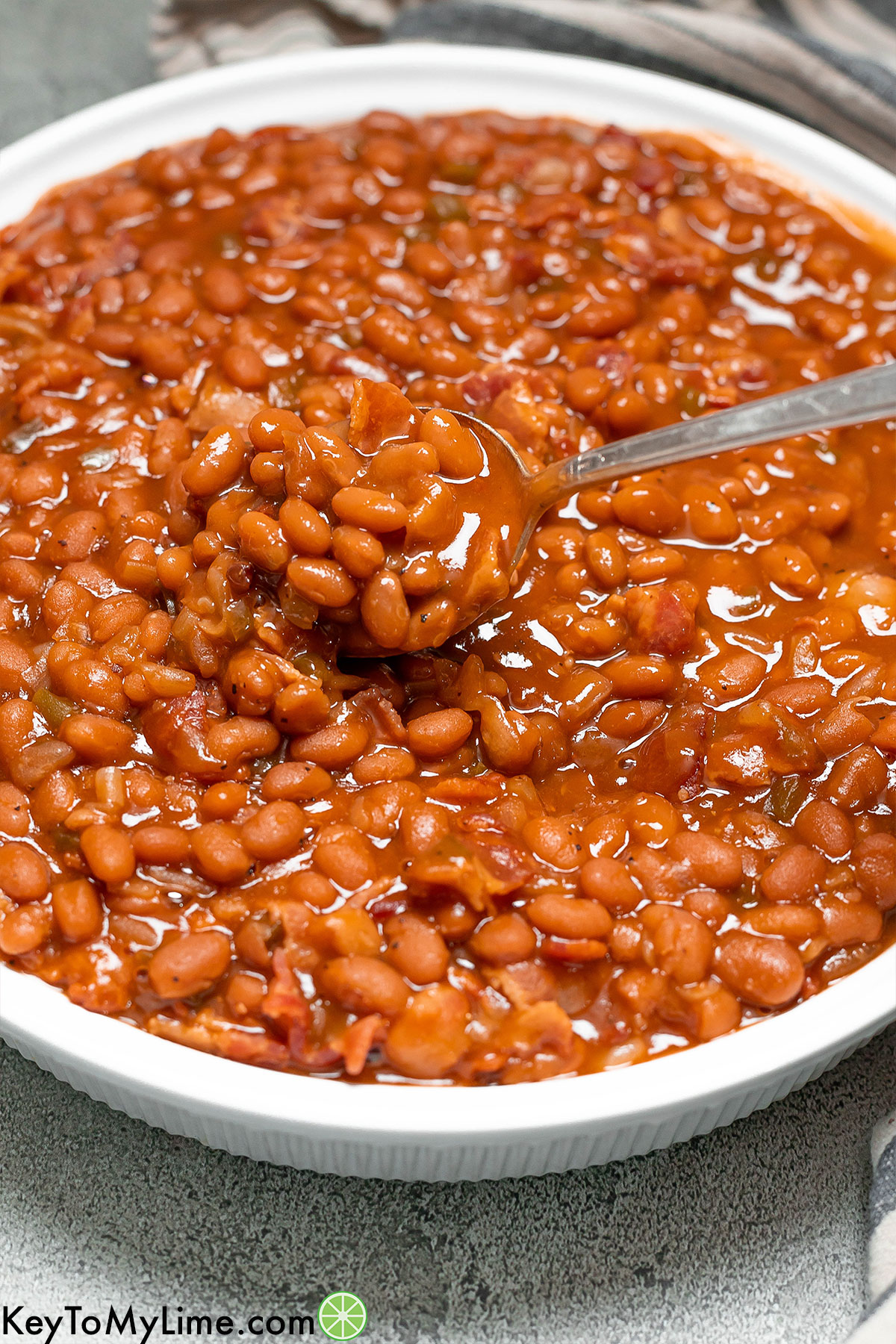 A serving spoon lifting baked beans up with green peppers and bacon pieces throughout.