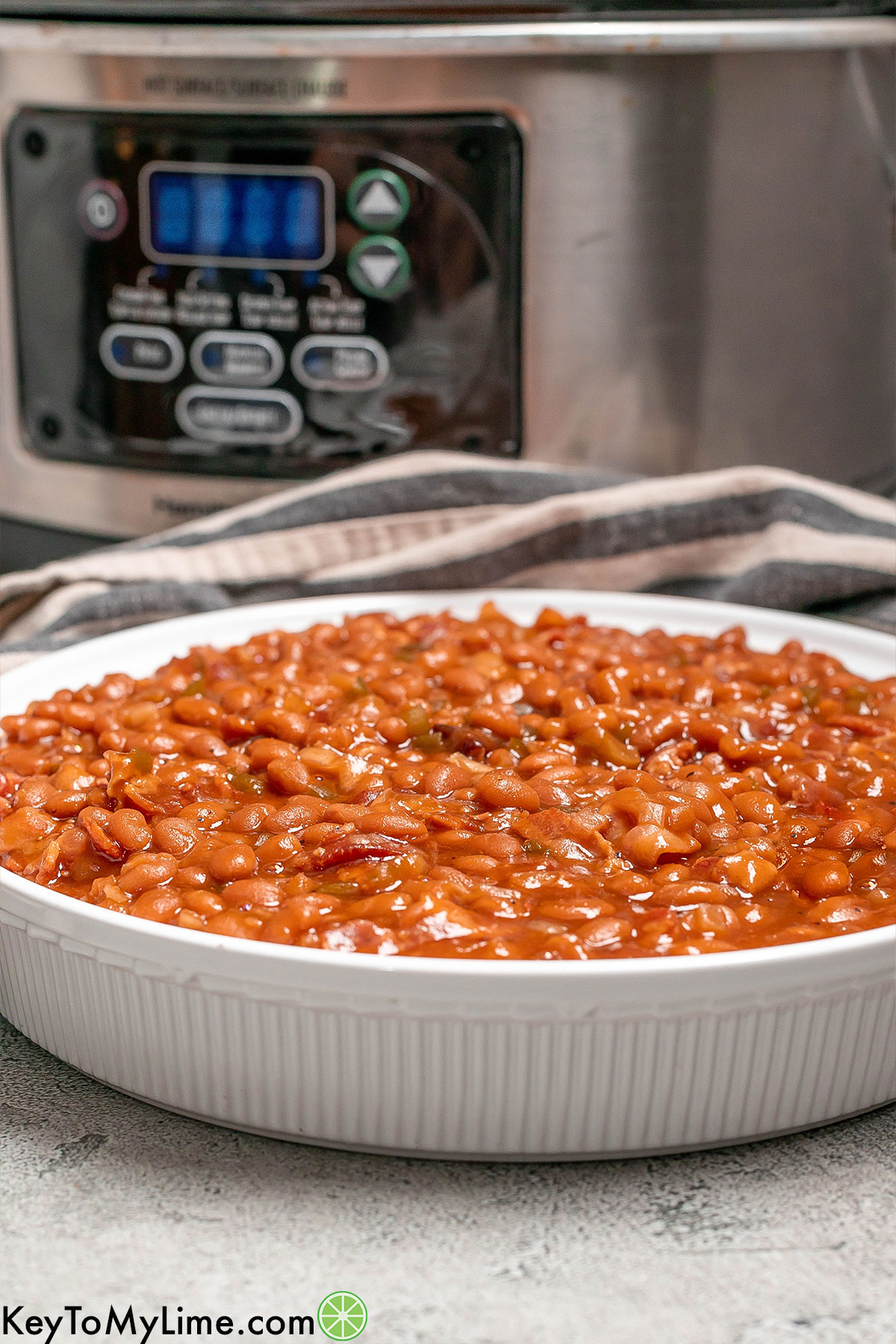A side image of a baked bean dish served in a white bowl with crockpot in the background.