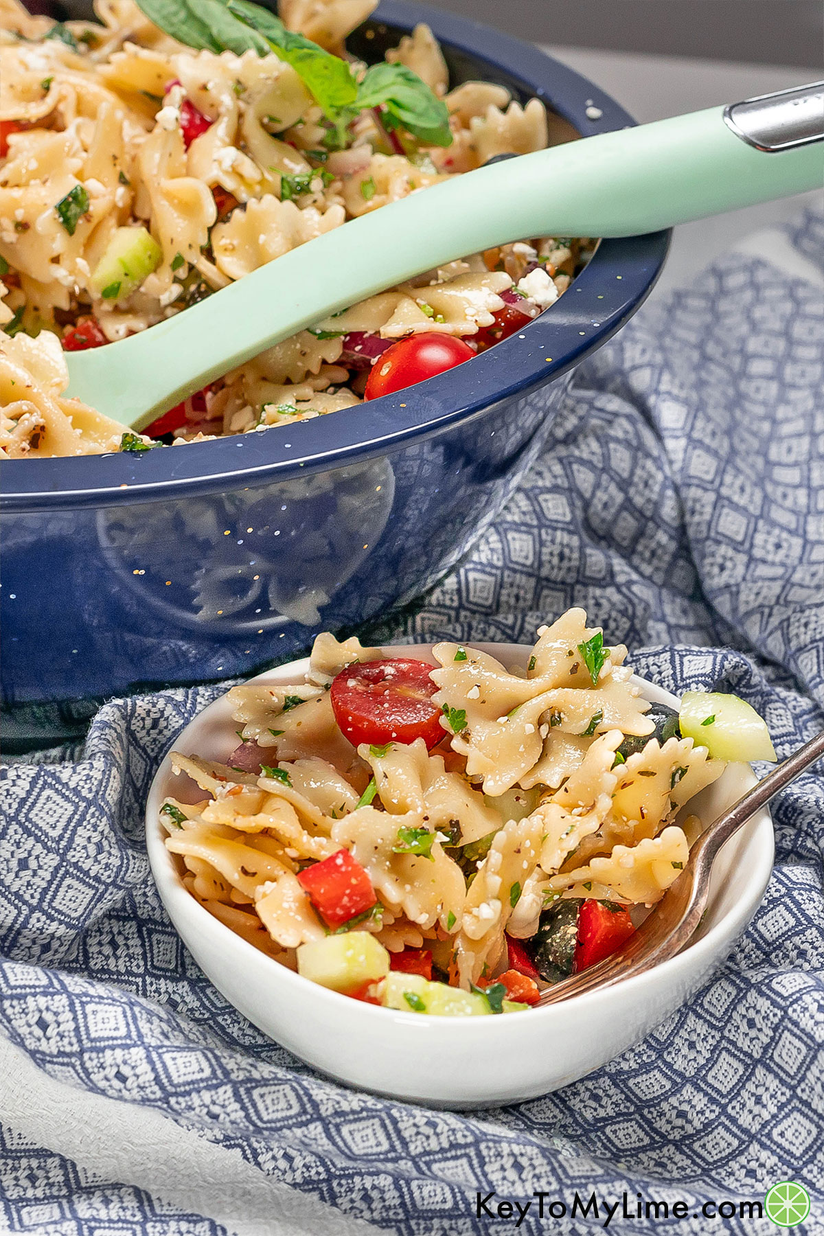 A side shot of pasta salad in a small serving bowl next to a large mixing bowl full of pasta salad.