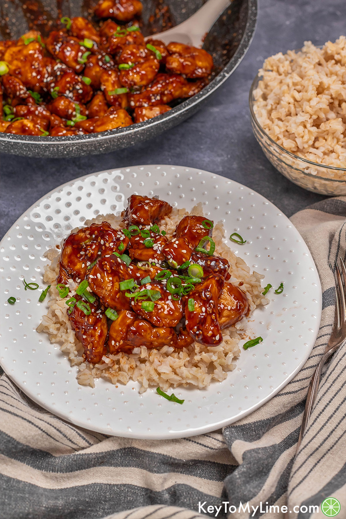 A plate of honey sticky chicken with a bowl of rice to the side and a pan in the background.