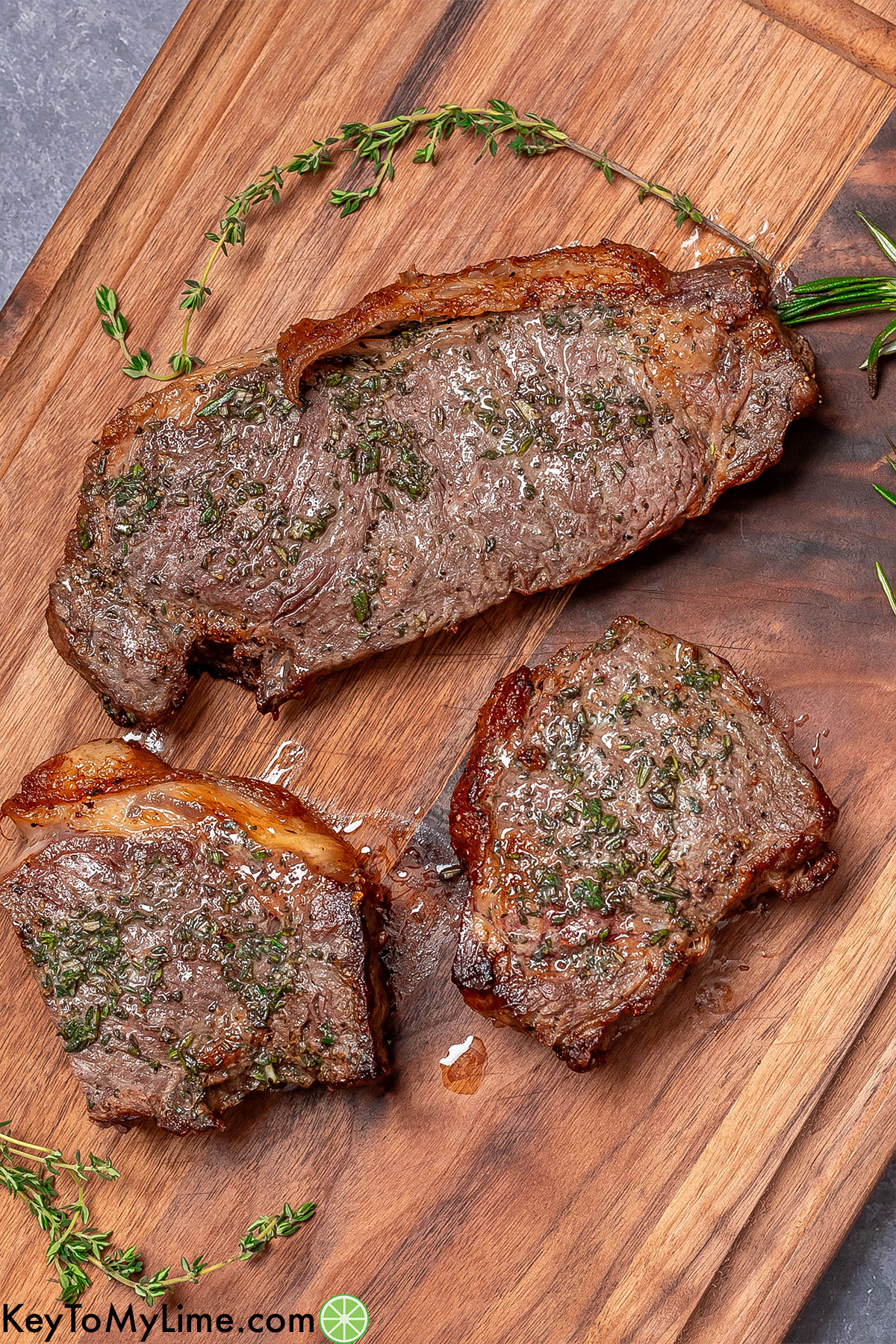 An overhead image of a few steaks resting on a cutting board with fresh herbs throughout.