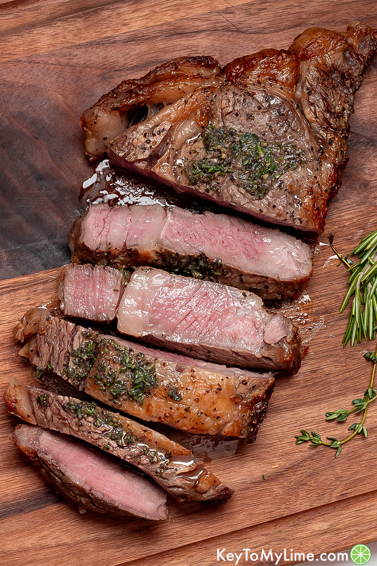 A sliced ribeye steak on top of a wood cutting board with fresh herbs throughout.