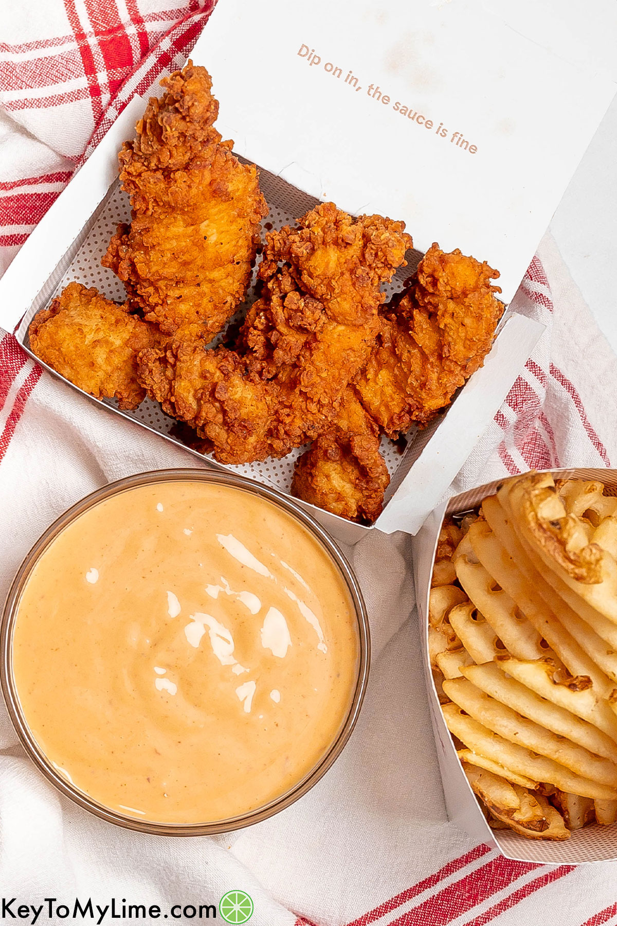An overhead image of two white boxes filled with crispy chicken tenders and waffles fries next to a medium sized bowl of homemade Chick Fil A suace.