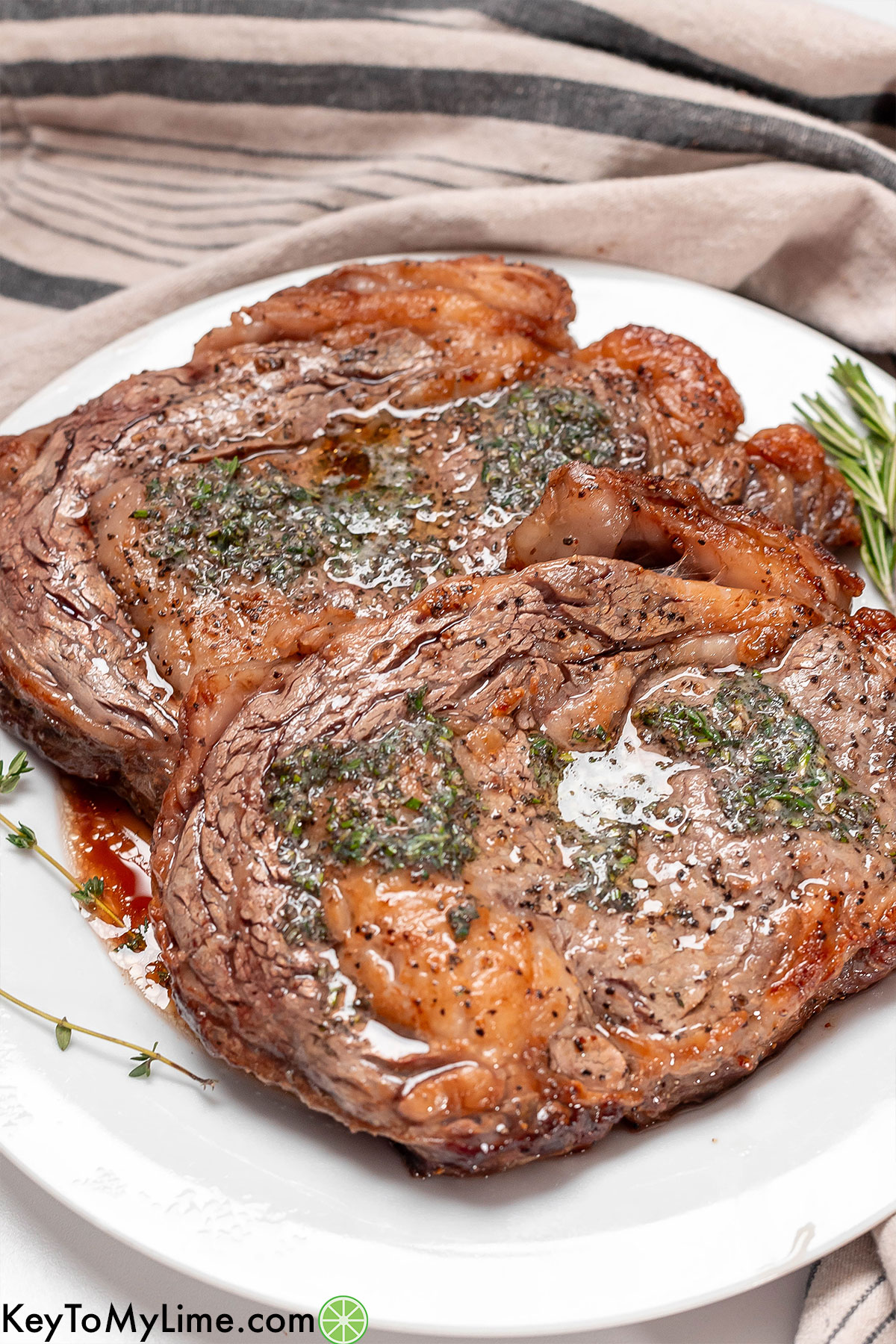 An angled image of a freshly cooked ribeye covered in fresh herb butter on top of a white plate.
