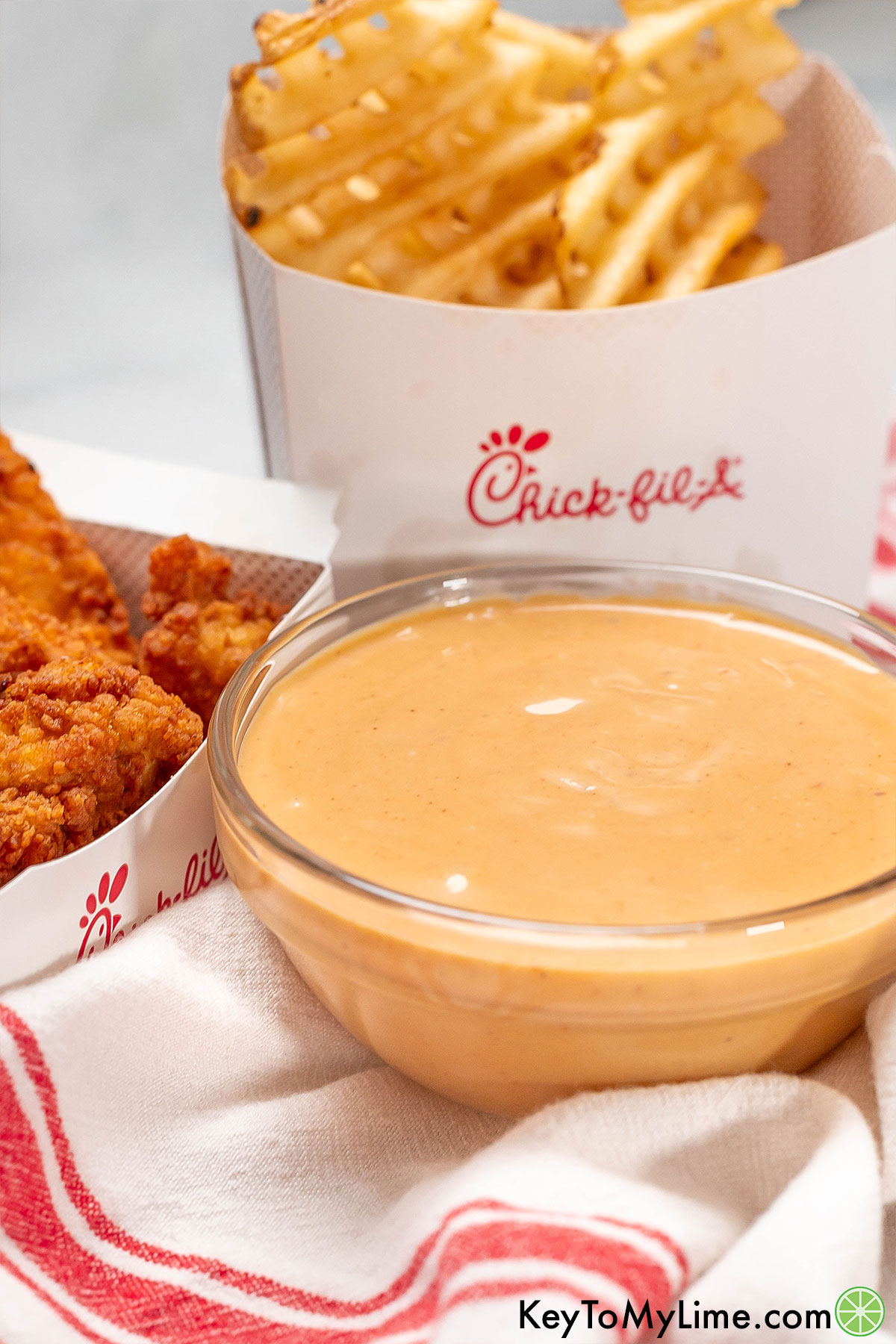 A side image of a medium sized glass bowl filled with freshly mixed sauce with a box of chicken tenders and waffle fries in the background.
