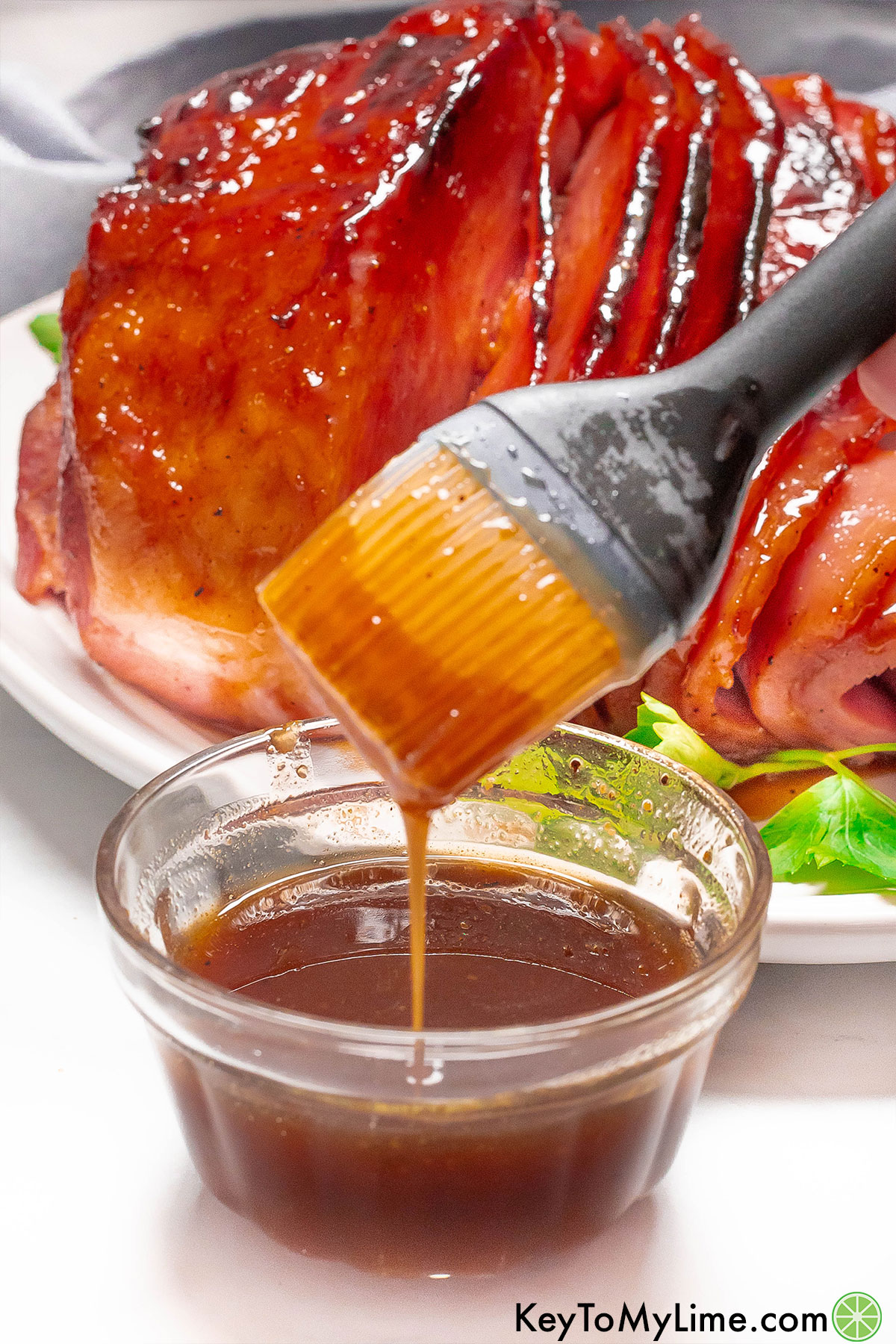 Freshly made ham glaze dripping from a basting brush with a cooked ham in the background.