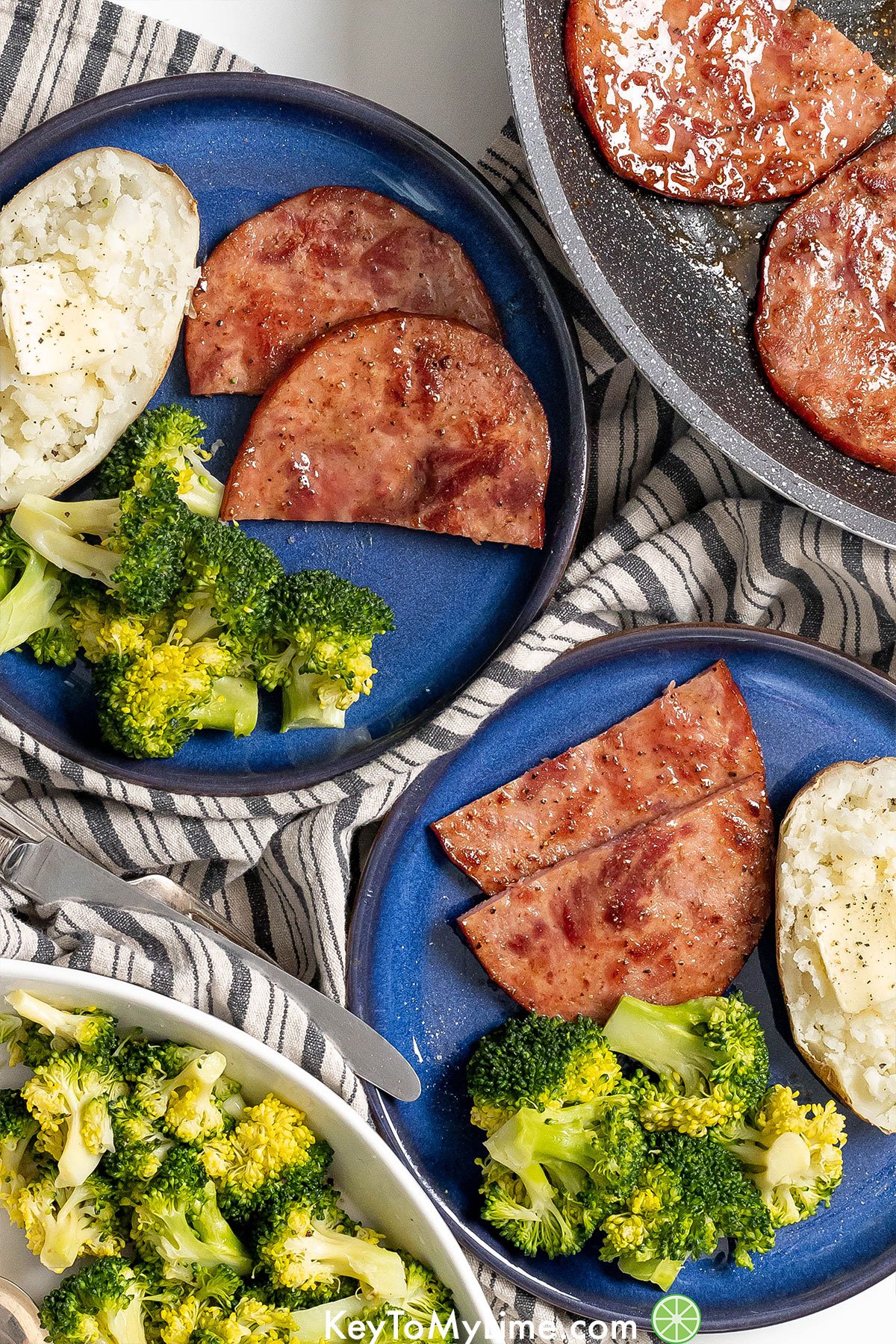 A couple of large dinner plates with ham steak, steamed broccoli, and a baked potato on top of a napkin.