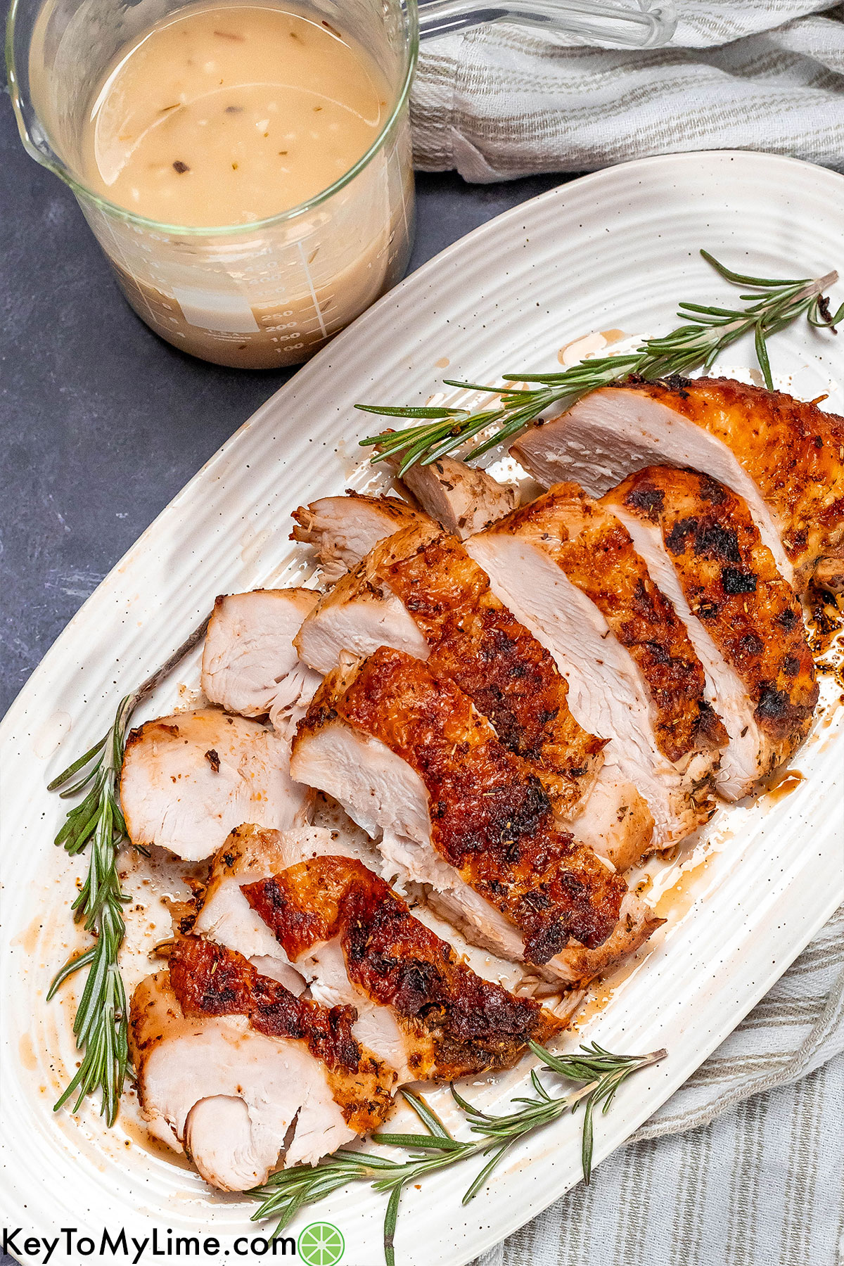 An overhead image of a sliced turkey breast on a large white platter with a small container of gravy to the side.