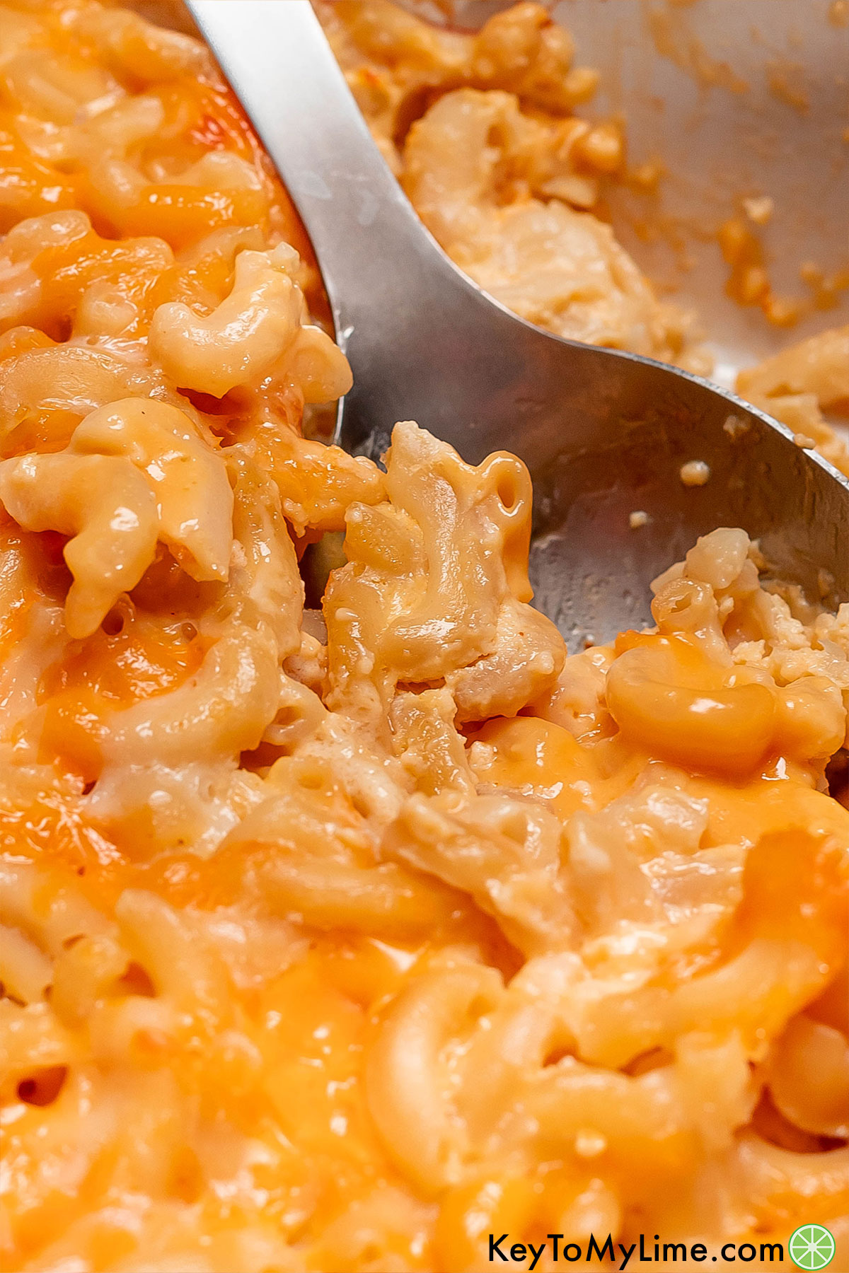 A close up image of a serving spoon lifting up over the rainbow mac and cheese in a casserole dish.