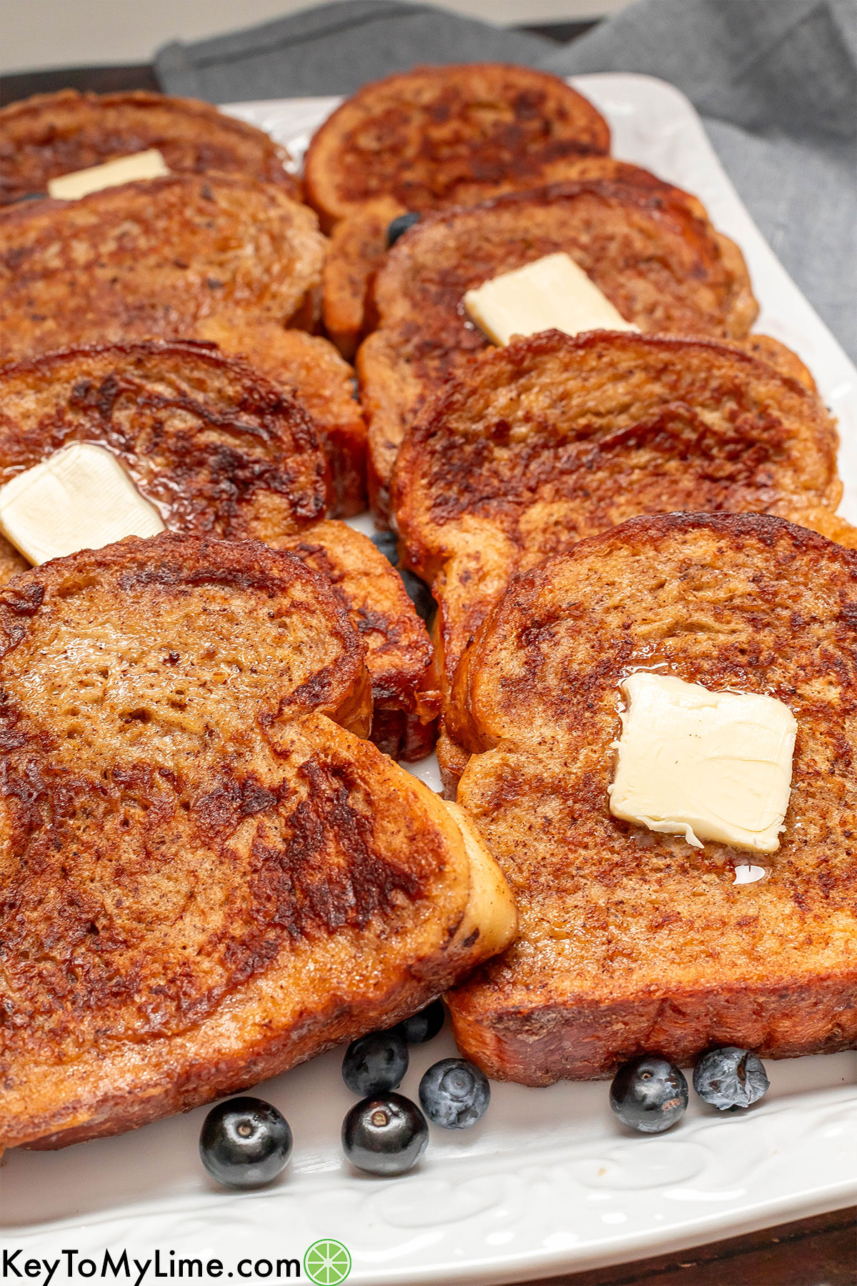 A side image of cinnamon French toast slices on a white platter with slabs of melting butter on top.