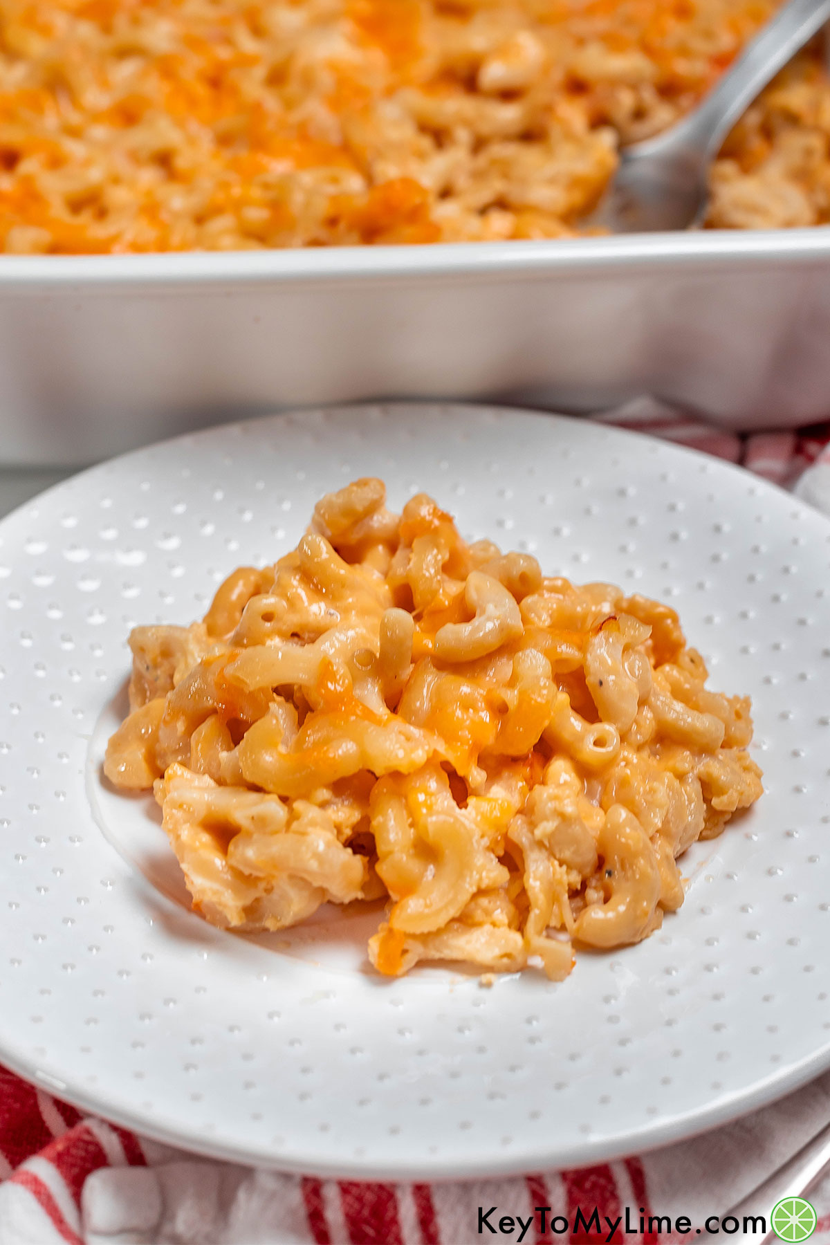 A side image of a serving of creamy mac and cheese on a white plate with a casserole dish in the background.