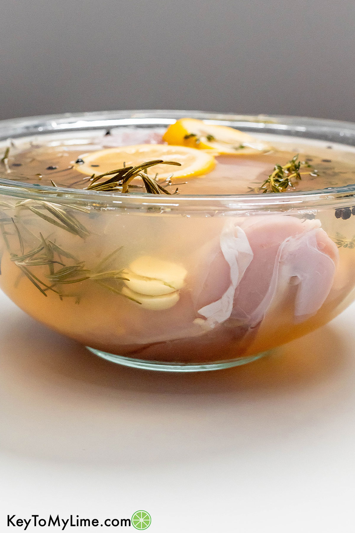 A side image of a turkey breast in brine with garlic, lemon wedges, and fresh herbs throughout in a large bowl.