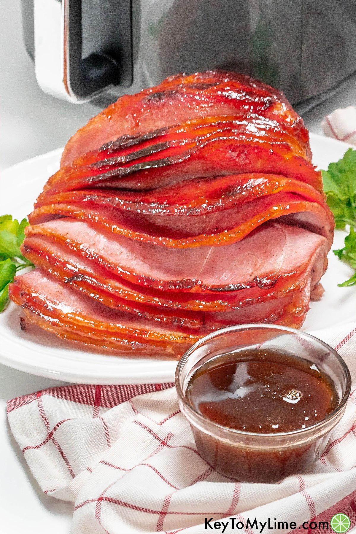 A side image of a spiral ham in front of an air fryer with a small bowl of ham glaze on top of a napkin.