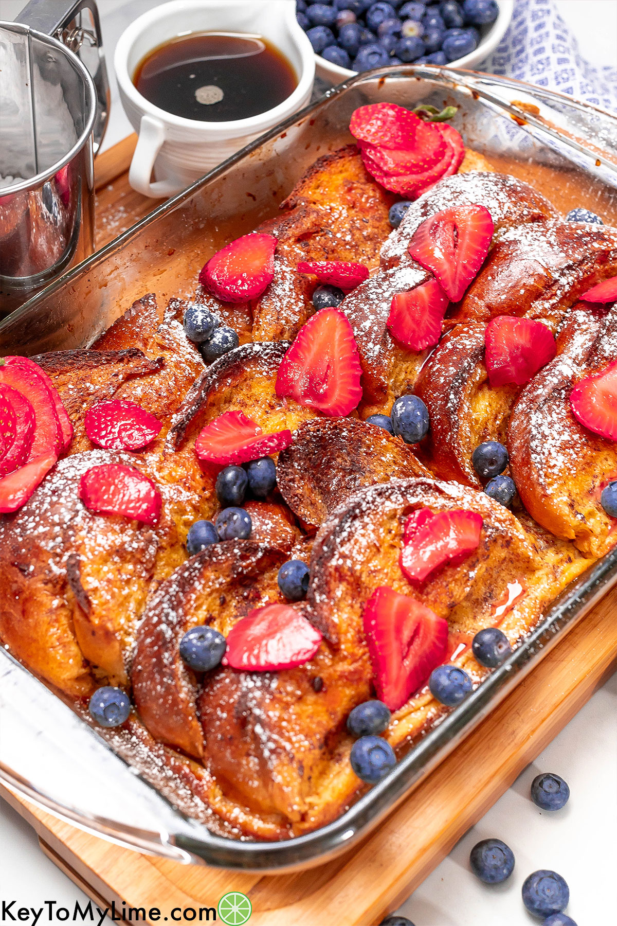 A whole freshly baked French toast casserole garnished with fresh berries, powdered sugar, and maple syrup.
