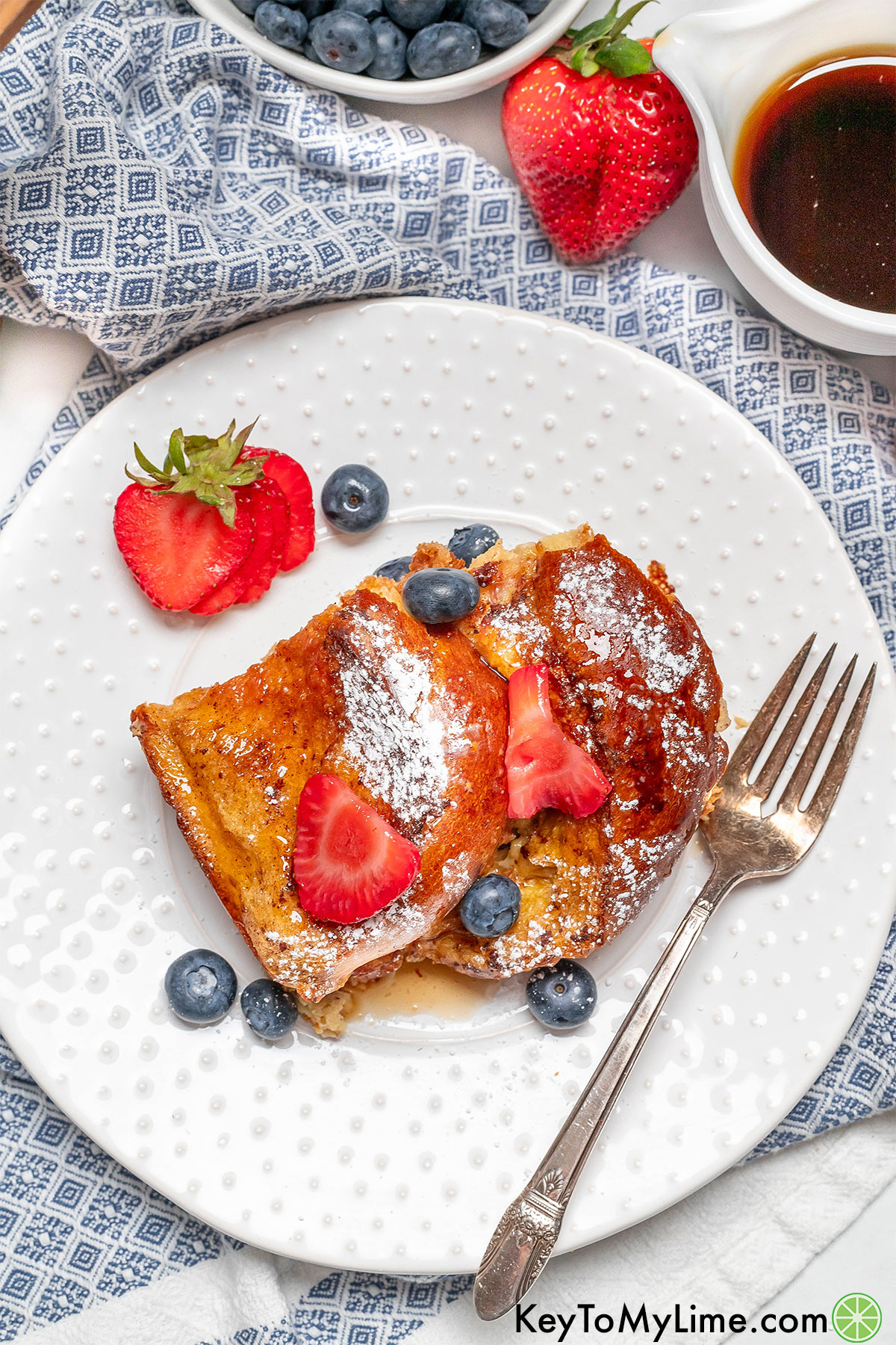 An overhead image of a beautifully garnished slice of French toast bake on a white plate on top of a colorful napkin.