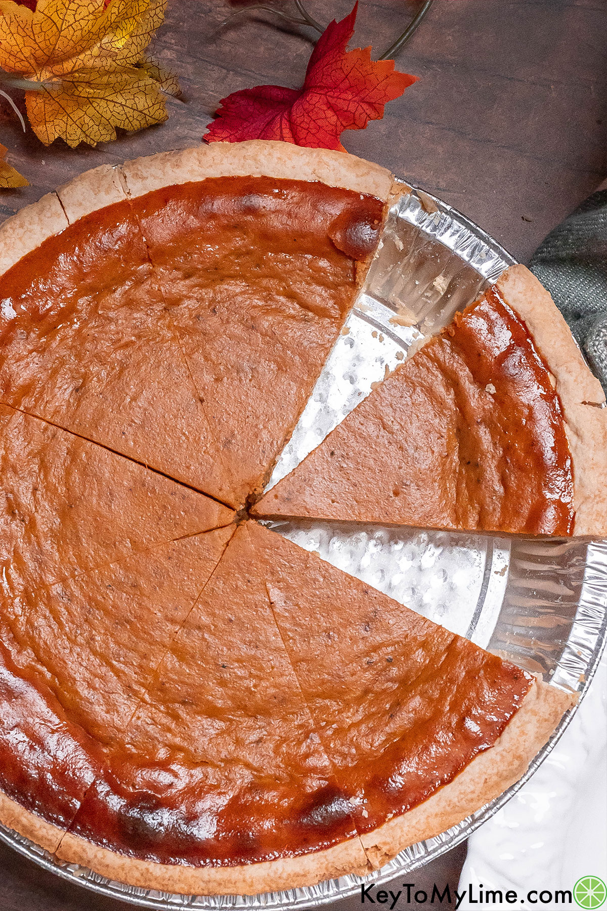 An overhead image of a pumpkin pie with slices missing and fall leaves around.