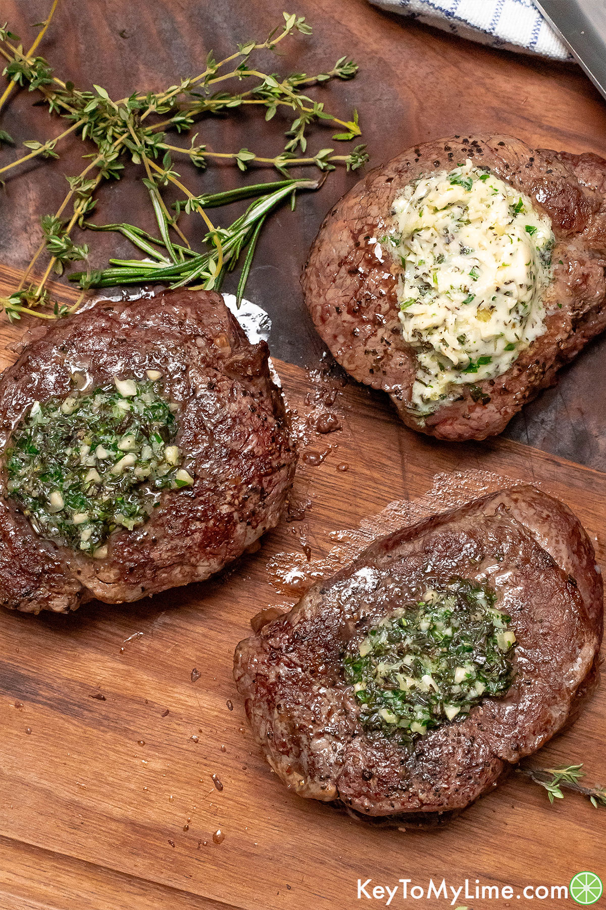 A group of three steaks with melted butter on top next to fresh herbs on a wood cutting board.