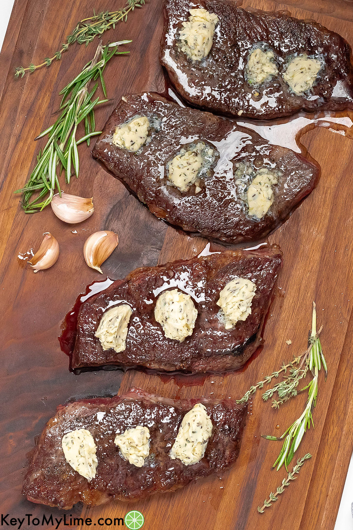 An overhead image of steaks resting on a cutting board with dollops of butter and herbs to the side.