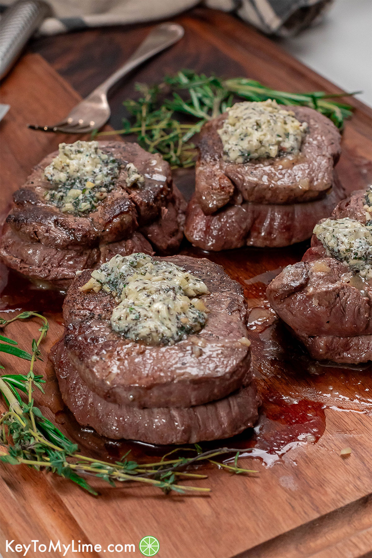 A side image of multiple filet mignons with herbs to the side resting on a cutting board.