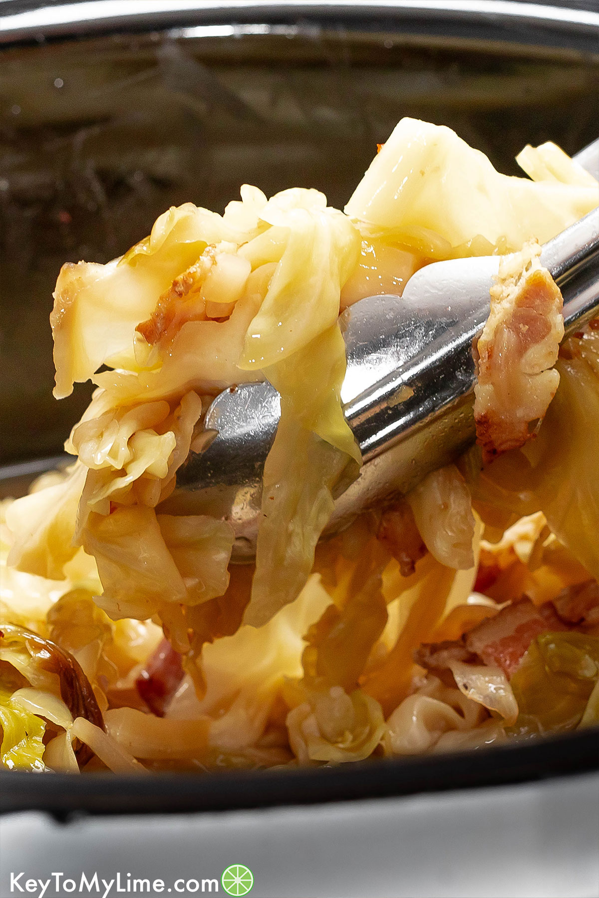Tongs lifting out of a crockpot with a scoop of cabbage and bacon.