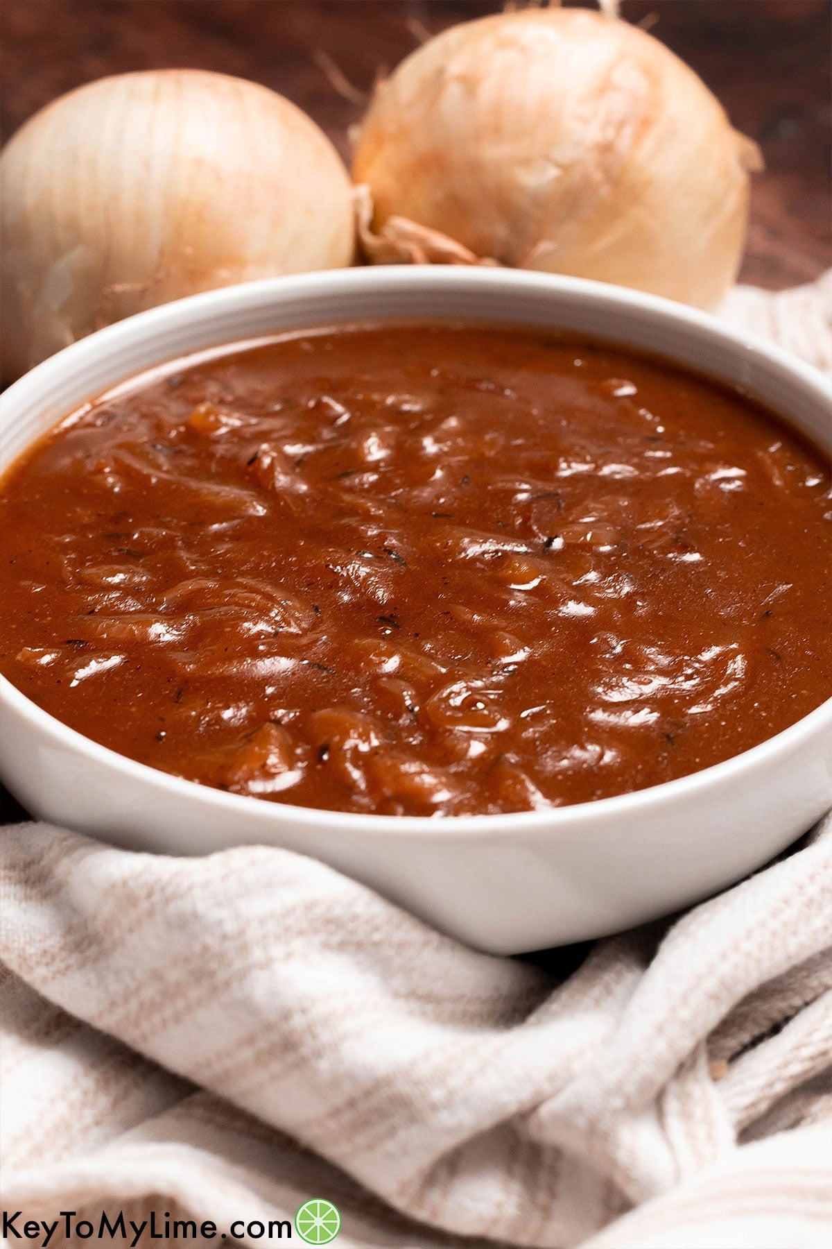 A side image of thick onion gravy in a white bowl with whole onions in the background.