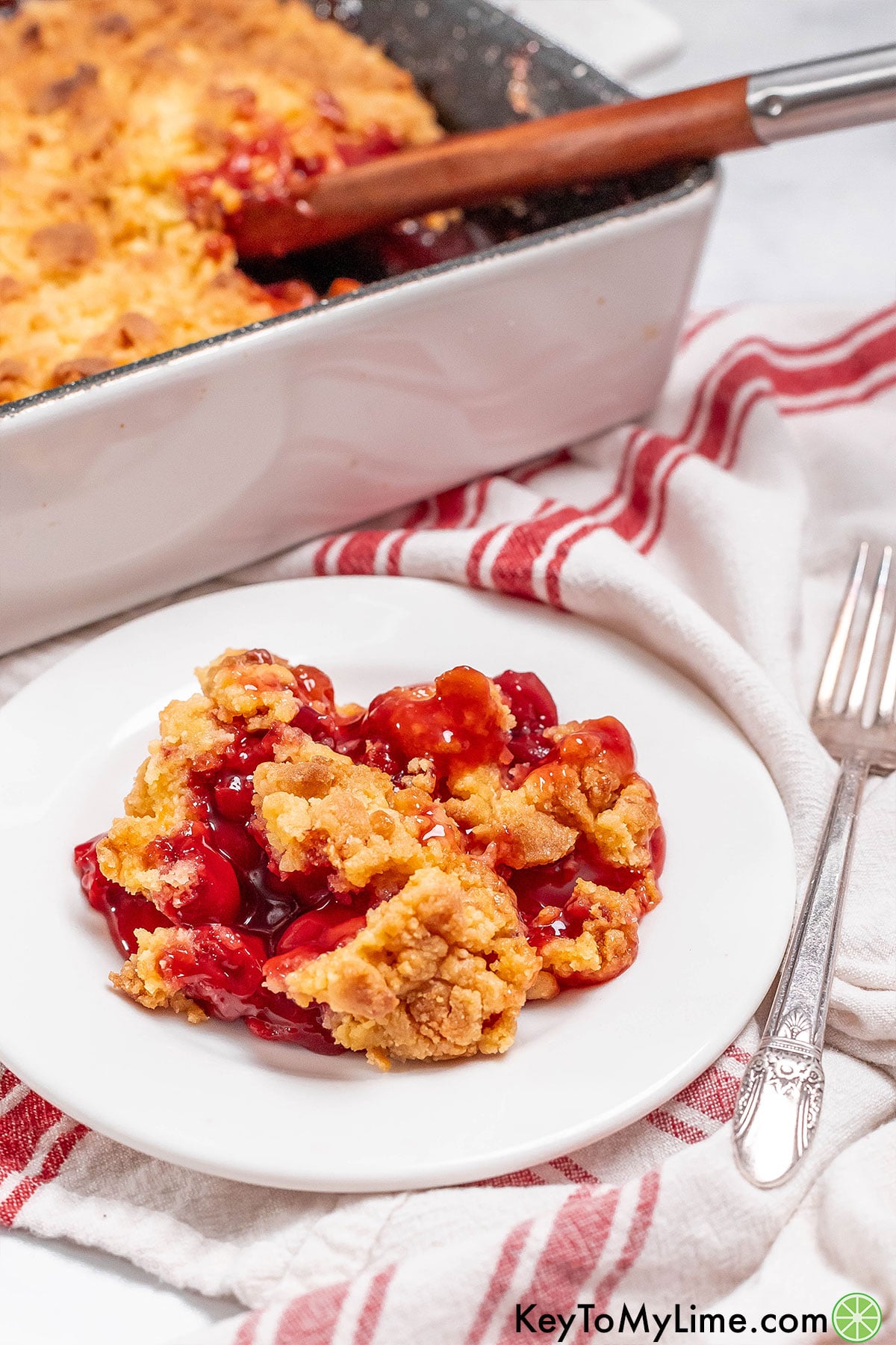 A side image of a serving of cherry cobbler with a partially filled baking dish in the background.